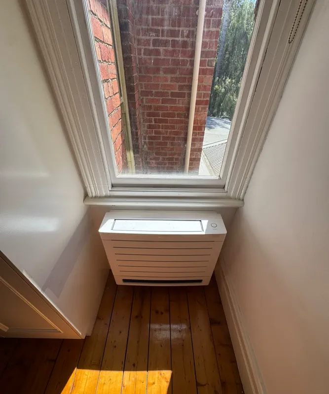 Floor-standing air conditioner under a tall sash window on polished floorboards