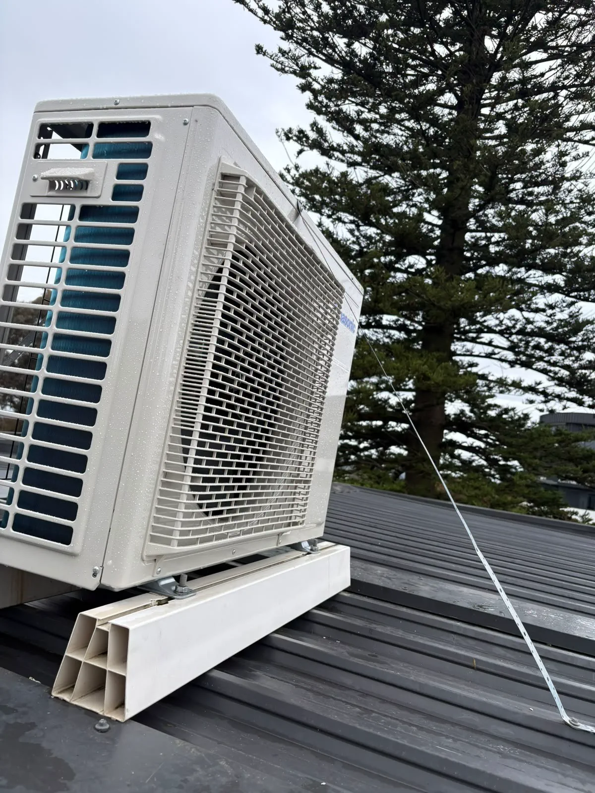 Split system outdoor unit on roof mounting blocks, strapped down to a metal roof