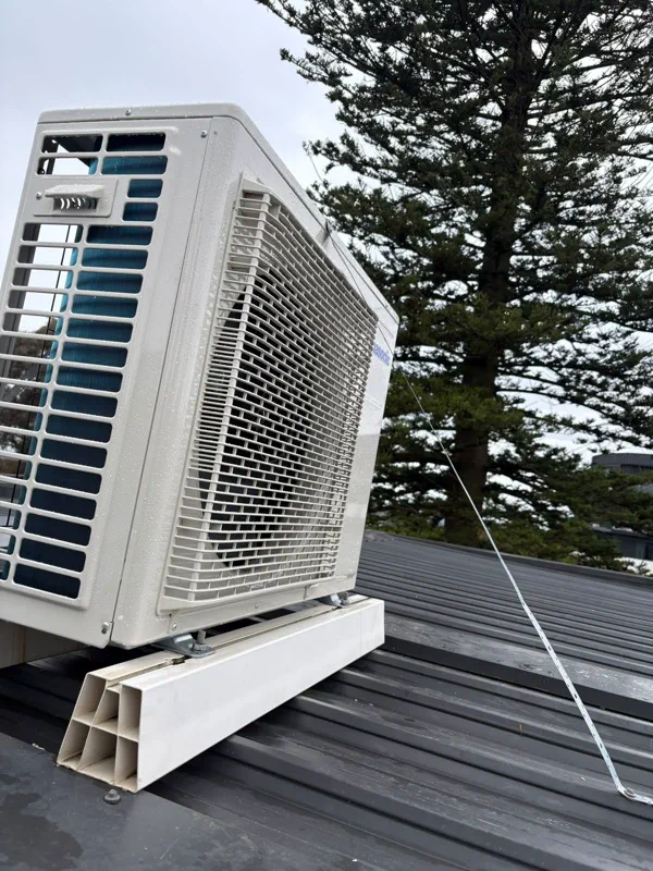 Split system outdoor unit on roof mounting blocks, strapped down to a metal roof