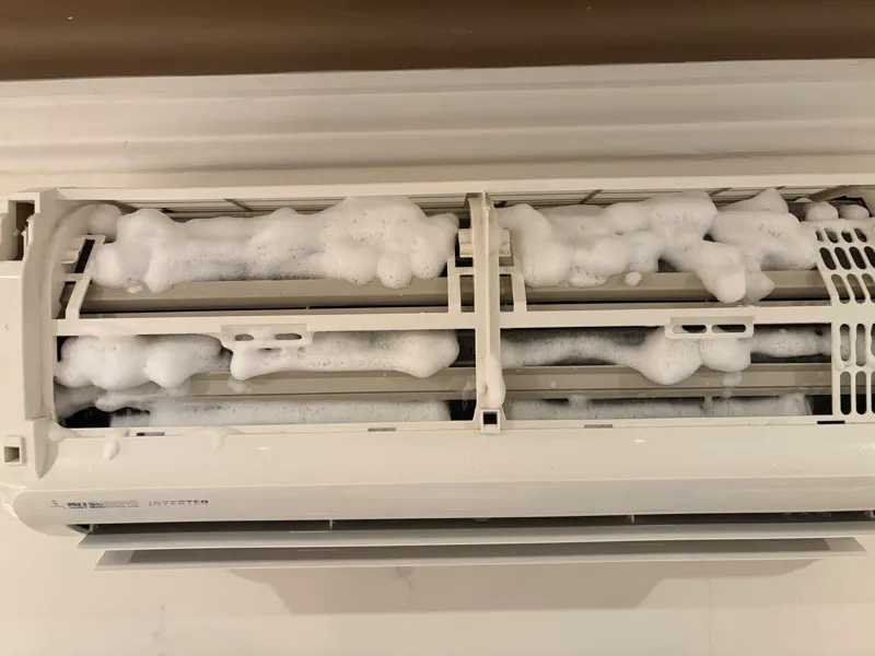 Wall split with the front grille lifted and cleaning foam across the coil