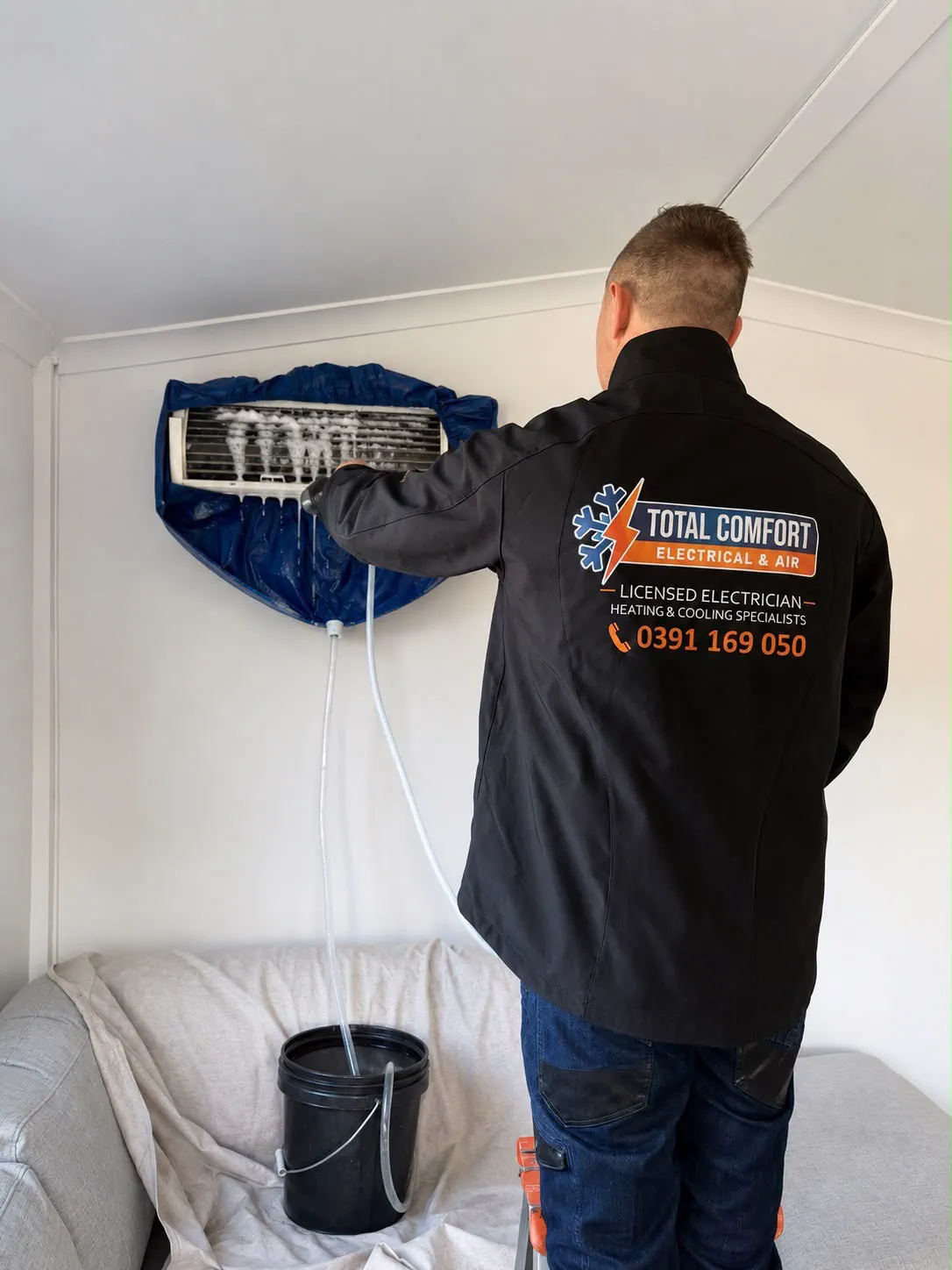 Total Comfort technician rinsing a split system coil inside a wash bag, with the couch below covered by a drop sheet