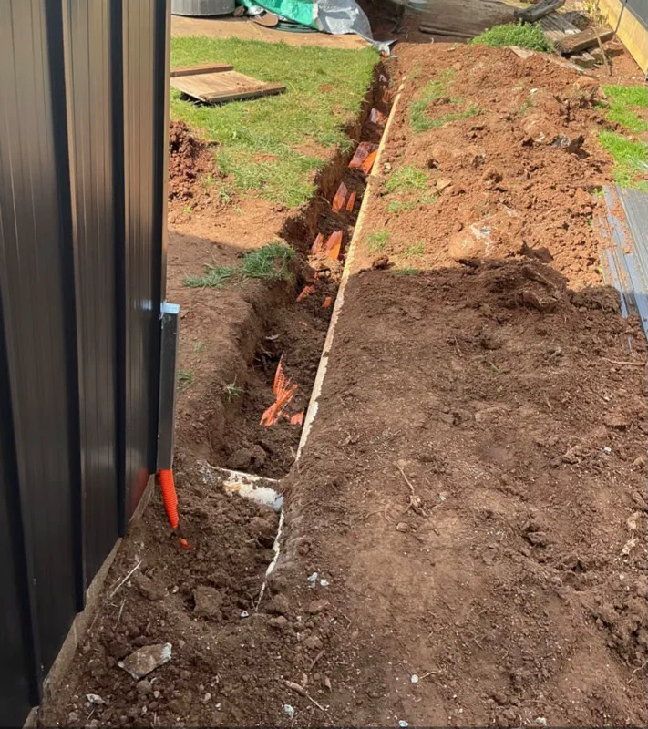 Open trench in a side yard with orange electrical conduit rising beside a fence post